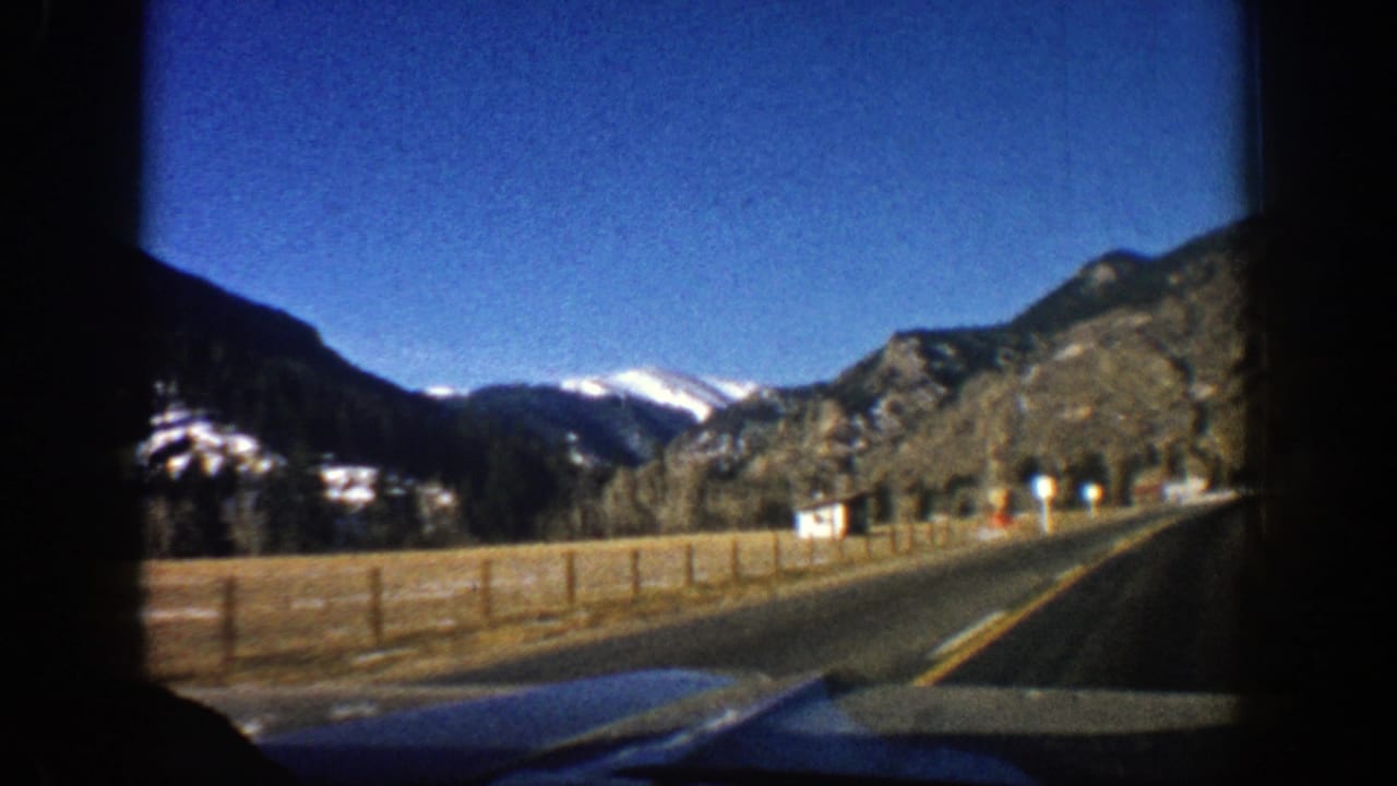 Mountain road seen from inside a car with fences, rocky slopes, and snow-capped peaks ahead.