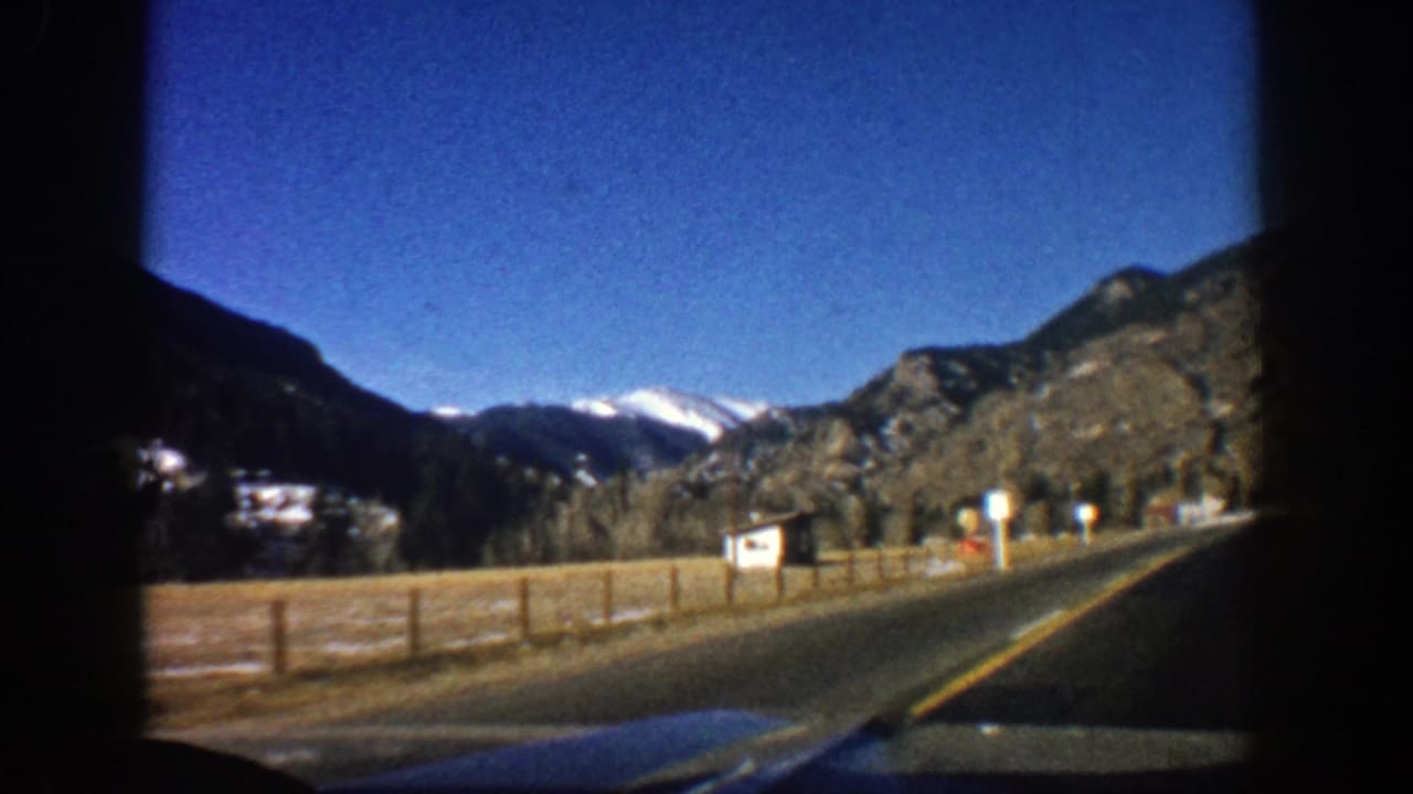 Fenced roadside curves toward rocky hills and distant snowy peaks under a blue sky.