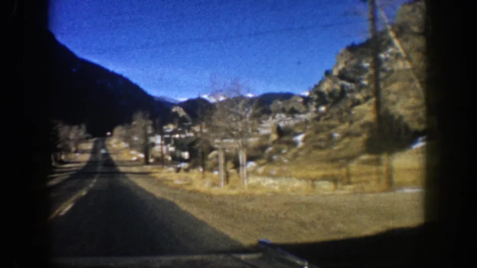 A brief 1961 Aspen car-window view down a mountain road with houses, fencing, rocky hills, and snowy peaks visible.