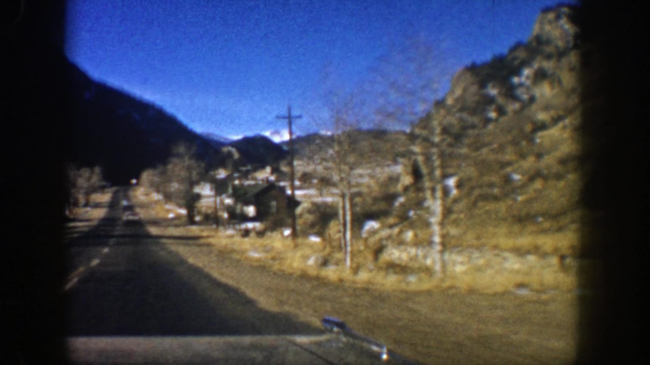1961 Aspen-area road view from a car, showing a paved road, rocky slope, utility poles, sparse trees, and a small roadside house.