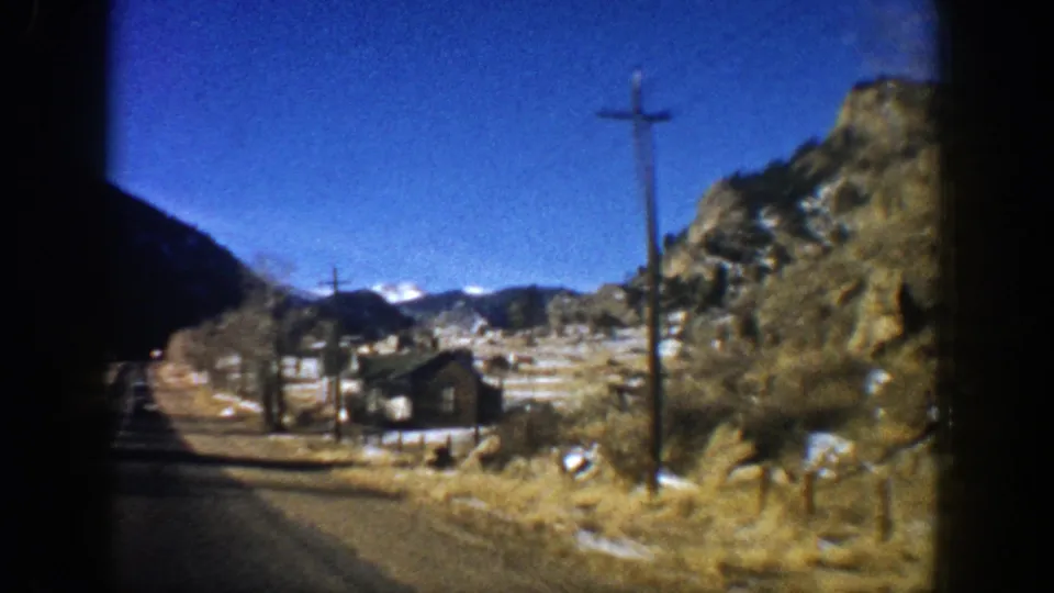 Aspen, Colorado home-movie frame showing a brown roadside house, utility poles, rocky hillside, and snow-dusted mountains beyond.