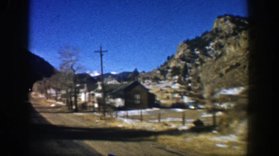 Closer 1961 Aspen roadside view with a small brown house, bare trees, utility pole, rocky hillside, and patchy snow under blue sky.