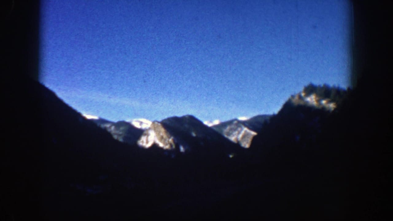 1961 Aspen, Colorado home-movie frame showing distant snow-capped mountain ridges beneath a blue sky and heavy shadowed foreground.