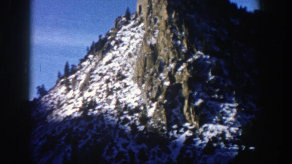 1961 Aspen, Colorado frame with a snow-dusted rocky mountain slope, scattered evergreen trees, and saturated blue sky.