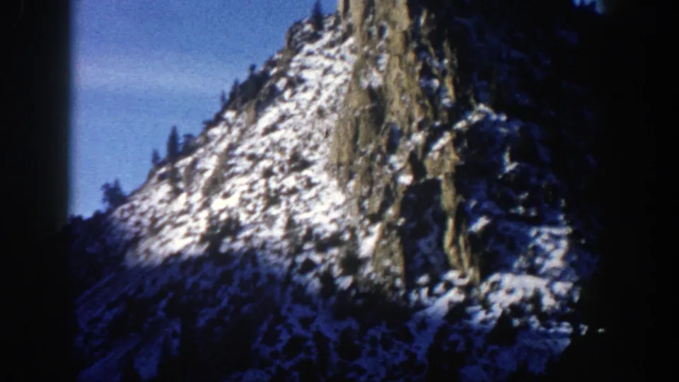 1961 Aspen, Colorado home-movie frame showing a steep rocky slope with patchy snow, dark trees, and soft 8mm grain.