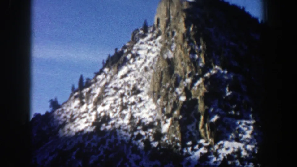 1961 Aspen, Colorado frame showing a snow-covered rock outcrop, dark evergreen treeline, and rounded 8mm frame edge.
