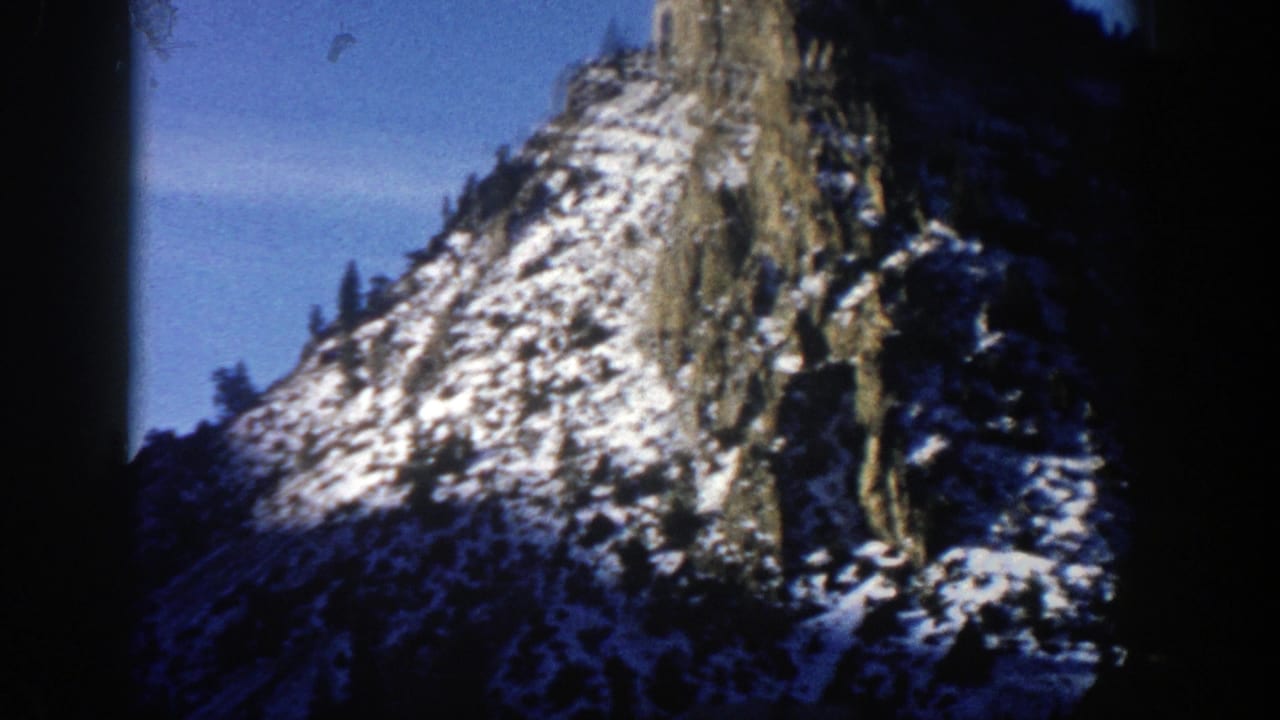 Snow-dusted rocky mountain peak under blue sky with dark wooded slopes below.