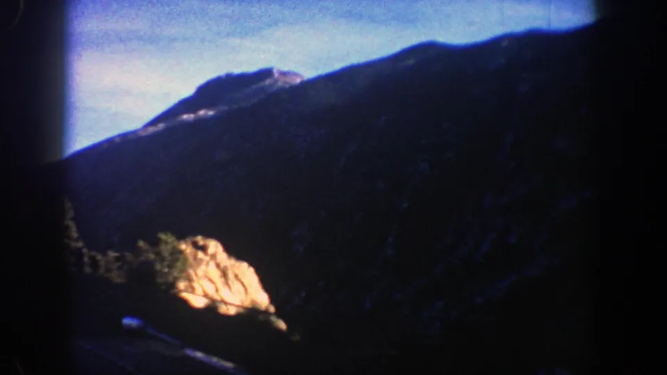 1961 Aspen-area 8mm frame showing a shadowed mountain slope, bright rock outcrop, evergreen trees, and blue sky.