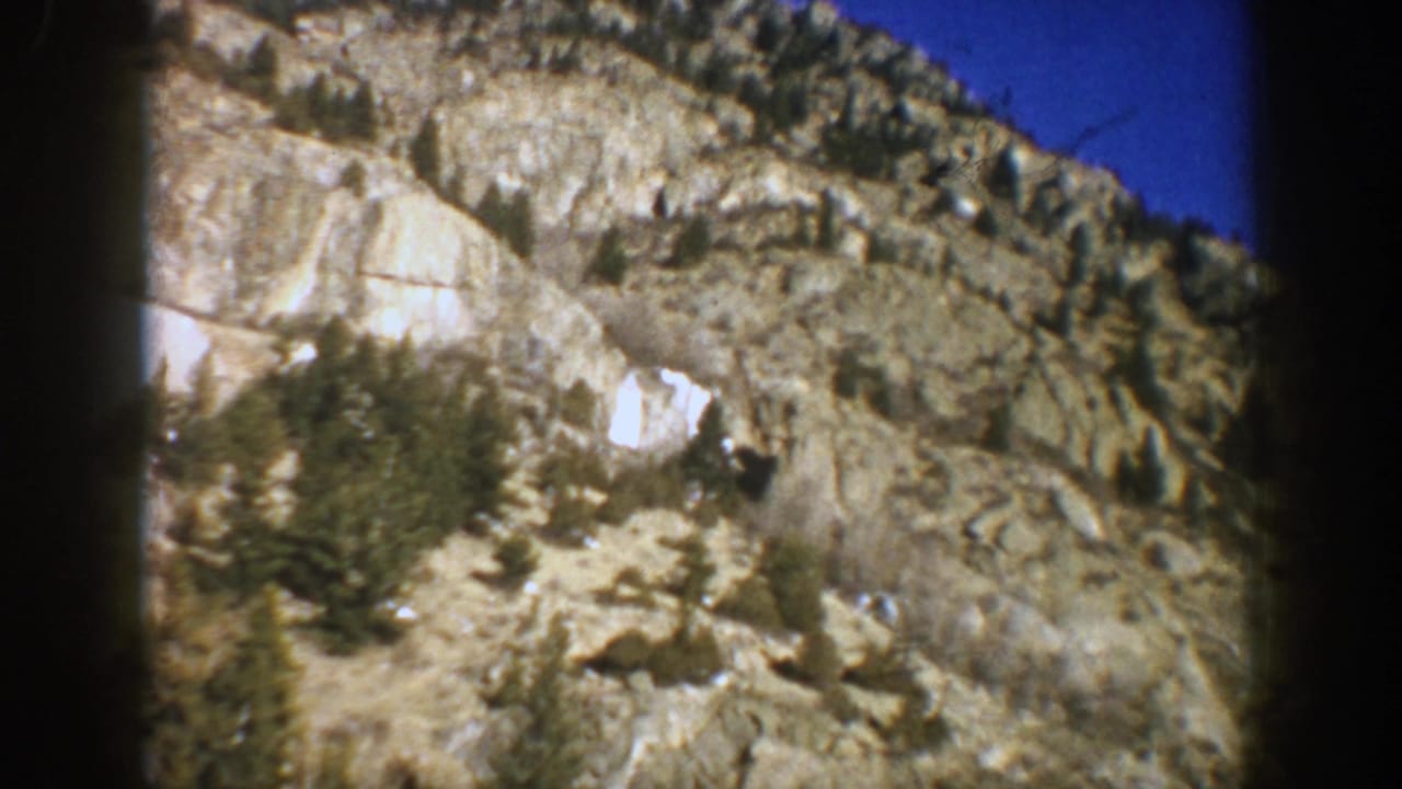 1961 Aspen, Colorado 8mm frame showing a sunlit rocky slope, scattered evergreens, and dark in-car framing.