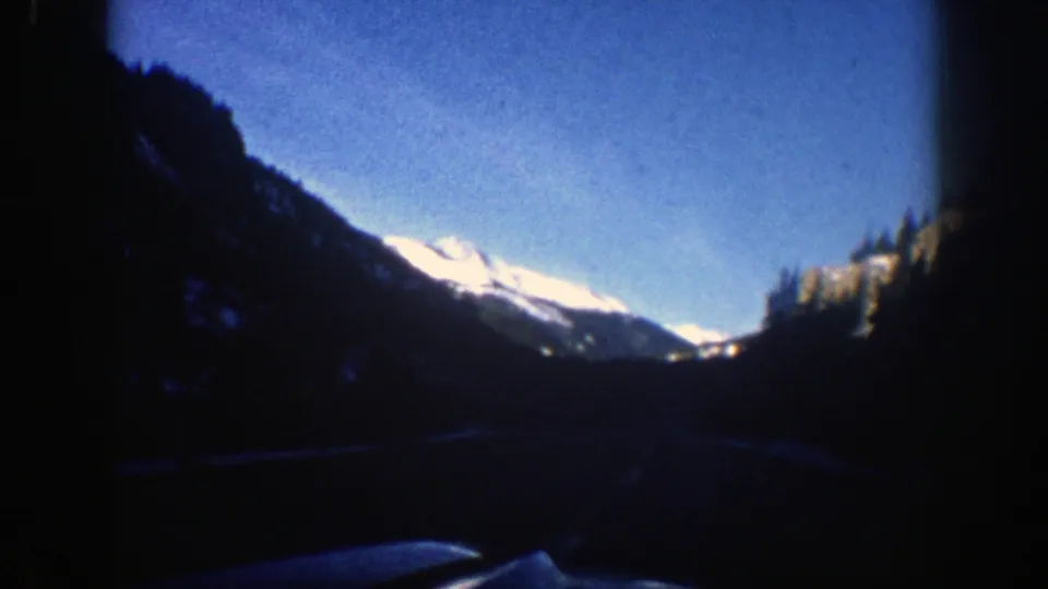Passenger-view 1961 Aspen road scene with dark dashboard framing and snow-bright mountains in the distance.