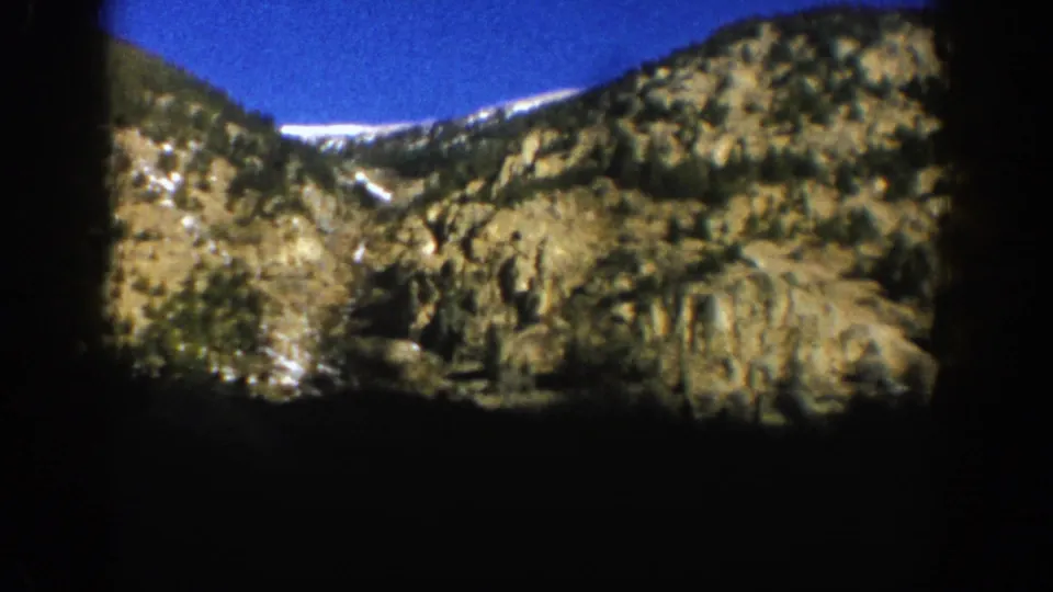 1961 Aspen 8mm view of rugged slopes, evergreens, and lingering snow patches from a moving car.