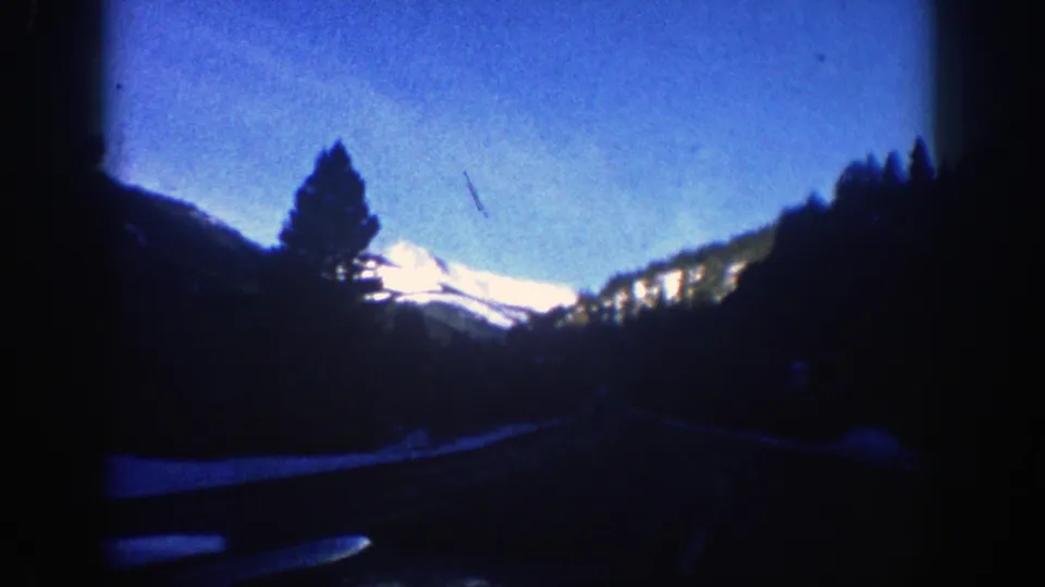 1961 Aspen, Colorado road scene with silhouetted evergreens, a curving mountain road, and bright snow-capped peaks beyond.