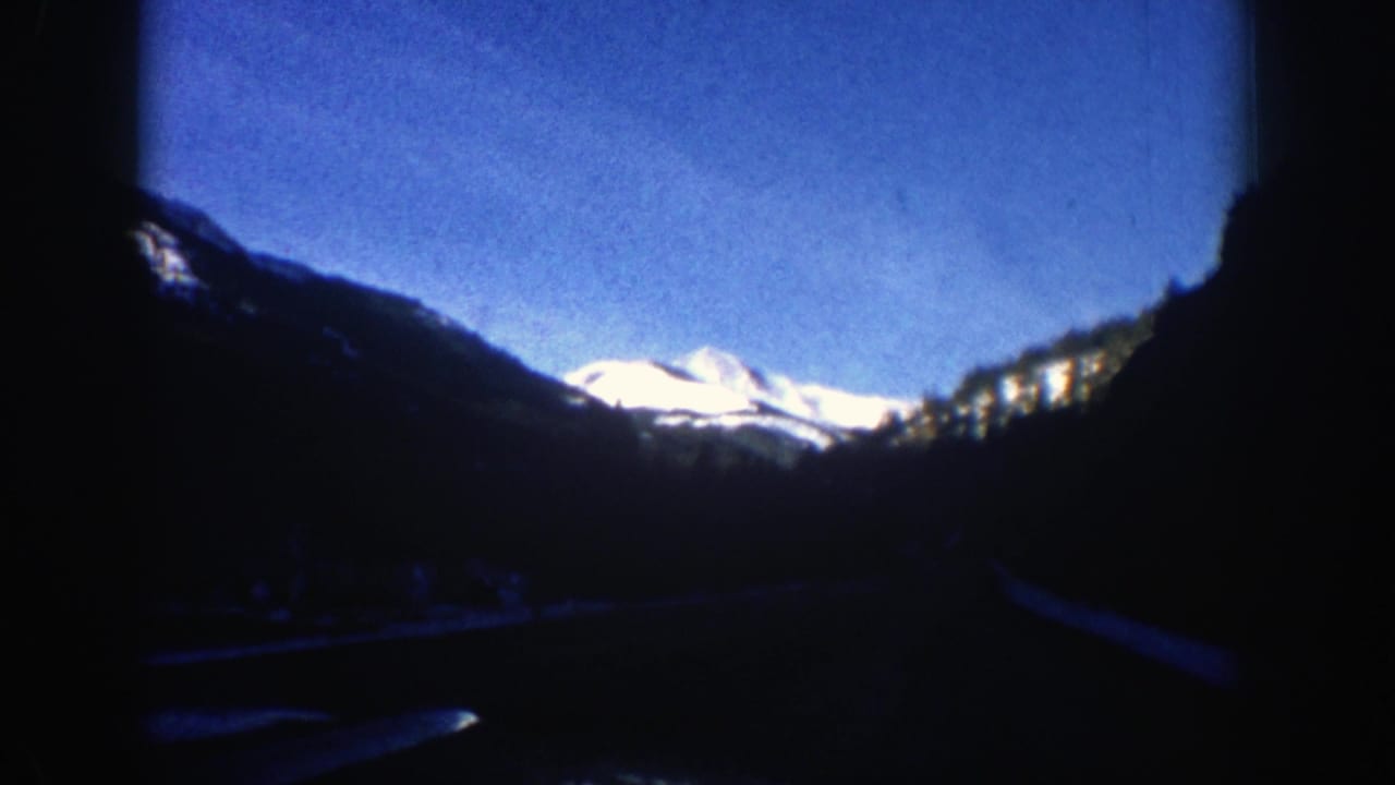 Snowy mountain peak beyond dark trees and a road seen from a moving vehicle.