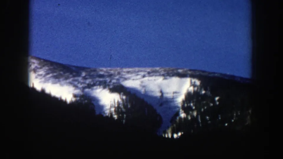 Aspen, Colorado 8mm frame showing a bright snow-covered slope, dark evergreens, and blue sky from a moving-car viewpoint.