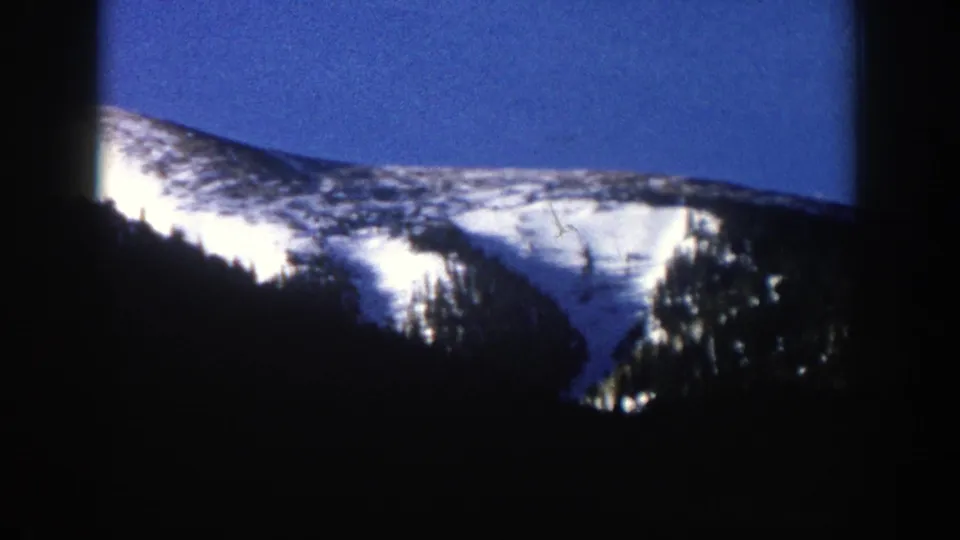 1961 Aspen, Colorado 8mm frame showing a sunlit snowy ridge behind shadowed evergreen slopes and a deep blue winter sky.