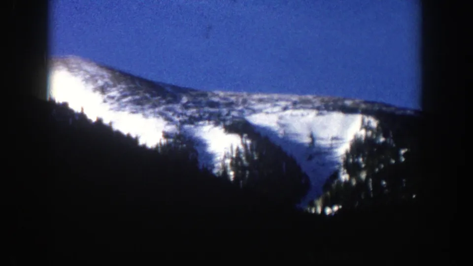 1961 Aspen, Colorado frame with a snow-covered ridgeline, shaded evergreen trees, and high-contrast winter light on 8mm film.