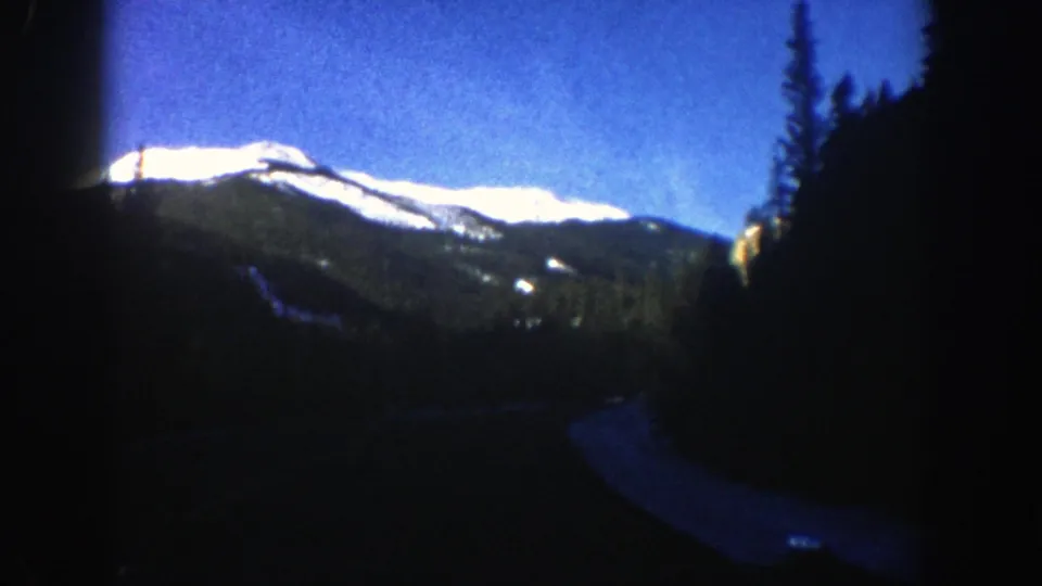 1961 Aspen, Colorado travel frame showing a curving road through a dark evergreen valley beneath distant snow-covered mountains.