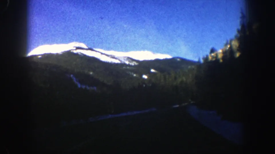 1961 Aspen, Colorado 8mm frame with a road curving through evergreen forest toward sunlit snow-covered mountain slopes.