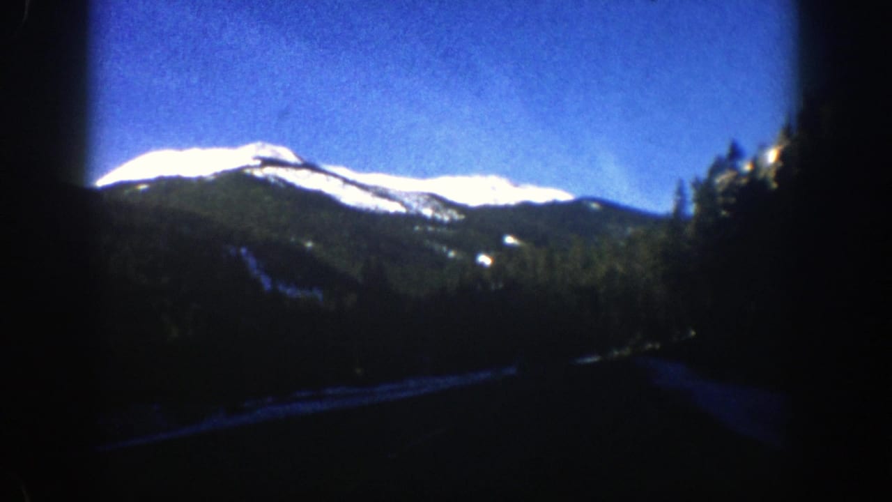 1961 Aspen-area mountain scenery viewed from inside a moving car, with snowy peaks, evergreen slopes, and a dark window edge.
