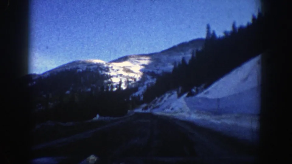 Aspen-area winter road seen from inside a moving car, with snowbanks, dark framing, and sunlit mountain slopes ahead.
