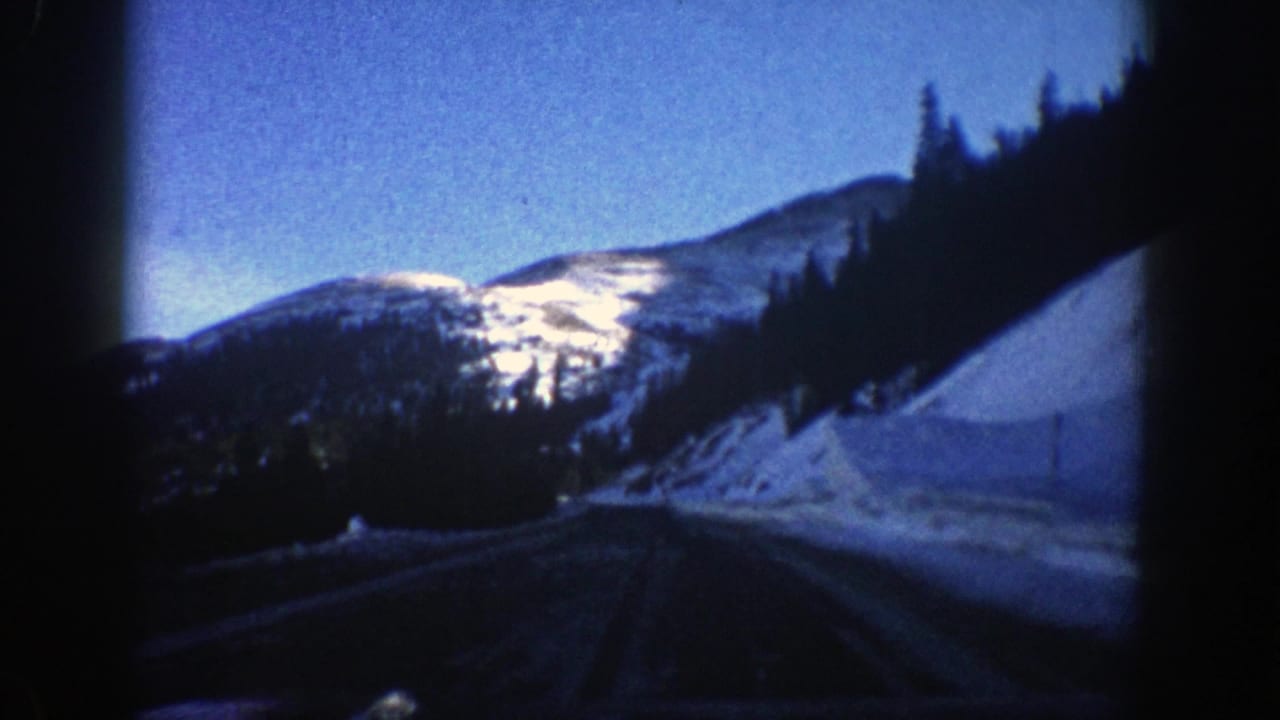 Snowy mountain road curving through evergreen forest under blue sky.