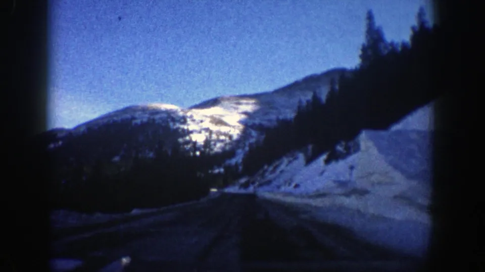 1961 Aspen-area road view from a moving vehicle, with snowbanks, evergreen hillside and a bright mountain ridge.