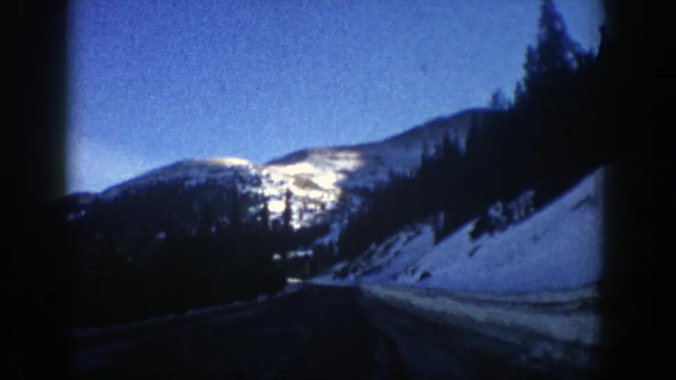 1961 Aspen-area 8mm frame with a dim windshield view of a road, snowbanks, evergreen trees and snowy mountain slopes.