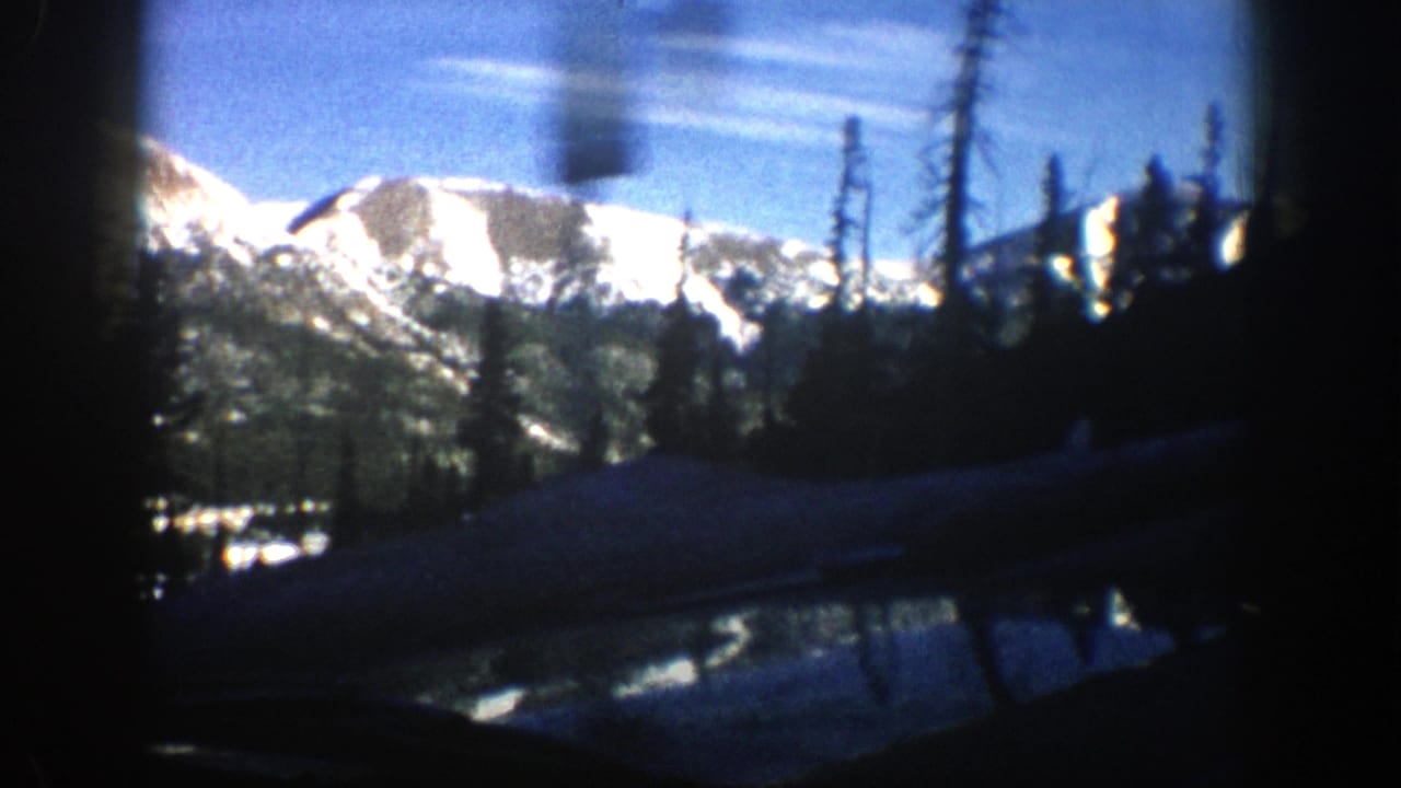 Snow-covered mountains and evergreens seen through a dark car-window frame.