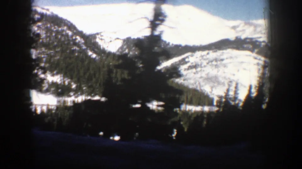 Aspen, Colorado mountain landscape with sunlit snowfields, evergreen hills, and dark tree silhouettes in the foreground.