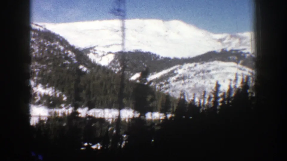 1961 home-movie frame of an Aspen-area snowy ridge and forested hillside seen from a moving roadside perspective.