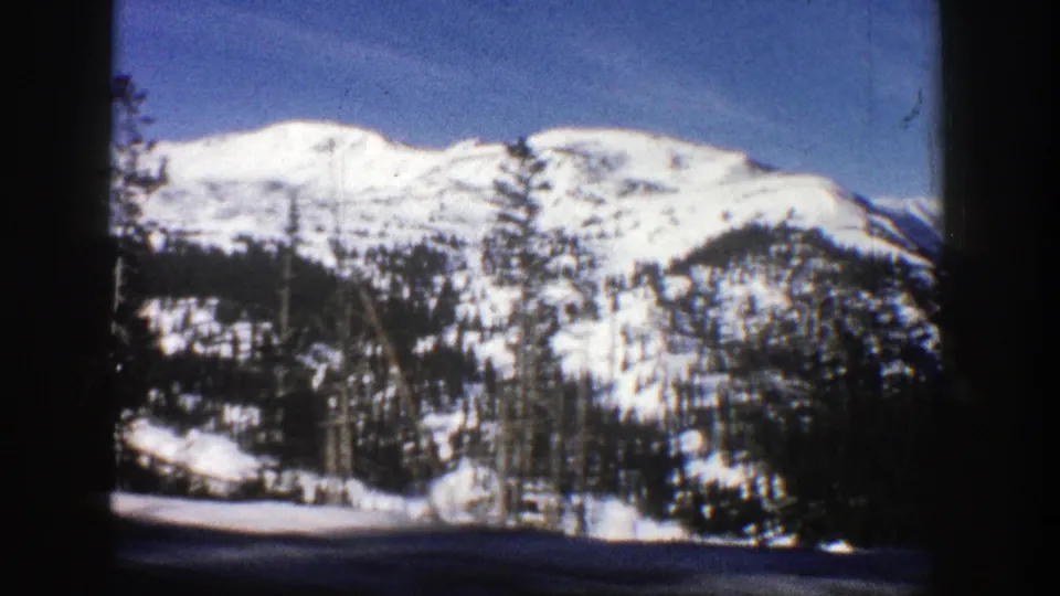 1961 Aspen, Colorado home-movie frame showing snowy mountain terrain, scattered evergreens, blue sky, and dark 8mm gate edges.