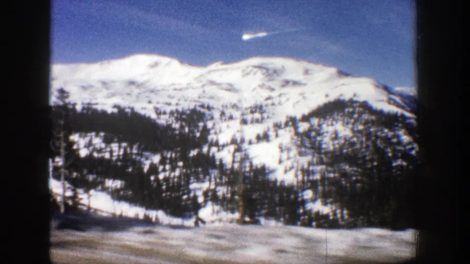 1961 Aspen, Colorado frame with a snow-bright mountain ridge, dark evergreen bands, blue sky, and small-gauge film borders.