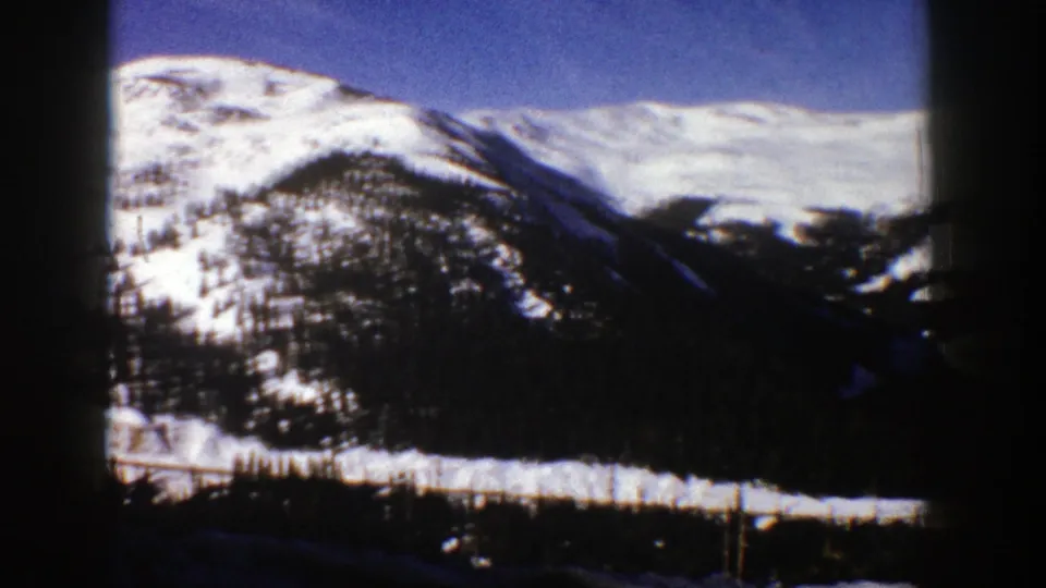 1961 Aspen, Colorado frame showing a snowy mountain valley, evergreen-covered slopes, blue sky, and dark foreground from the vehicle view.