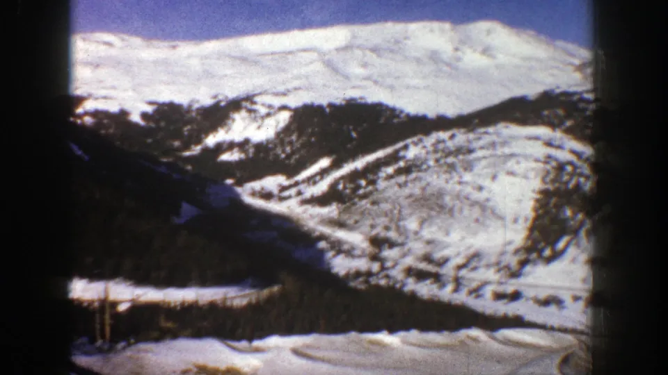 1961 Aspen, Colorado 8mm frame showing a rounded snowy hillside, darker tree-covered slopes, and distant winter ridges.