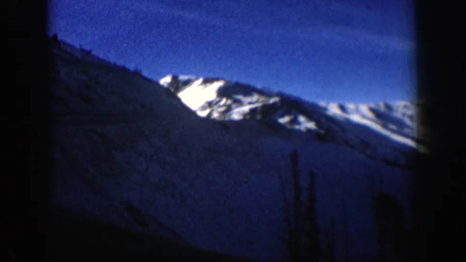 1961 Aspen, Colorado frame showing a shadowed slope in front of bright snow-covered mountain ridges and clear blue sky.