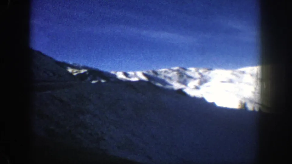 1961 Aspen, Colorado home-movie frame with bright snowfields across a ridge and a dark foreground hillside below blue sky.