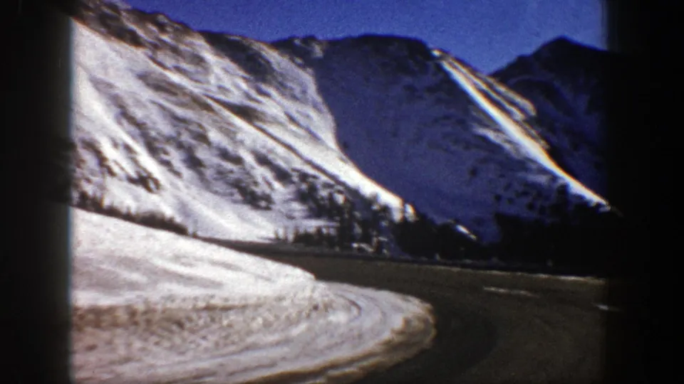 Aspen-area winter road view with a snowbank, dark road edge, evergreen trees, and steep Colorado mountain terrain.