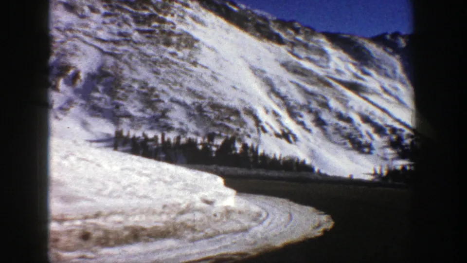 1961 8mm winter travel frame showing a road curve, snow-covered shoulder, evergreens, and rocky mountain slopes near Aspen.