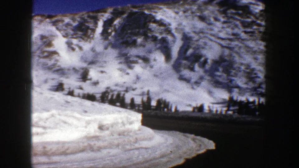 1961 Aspen-area 8mm frame showing a dark mountain road bending past snowbanks below snow-covered slopes.