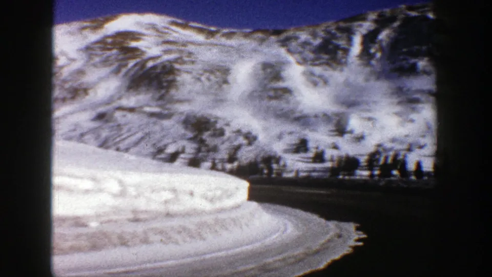 1961 Aspen, Colorado home-movie frame of a snow-lined mountain road curving under bright alpine slopes.