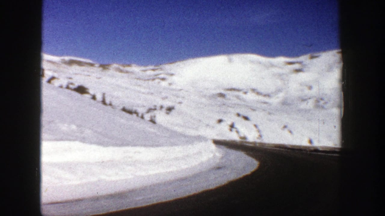 Snow-covered mountain slope with a curving road seen through dark vehicle-window edges.