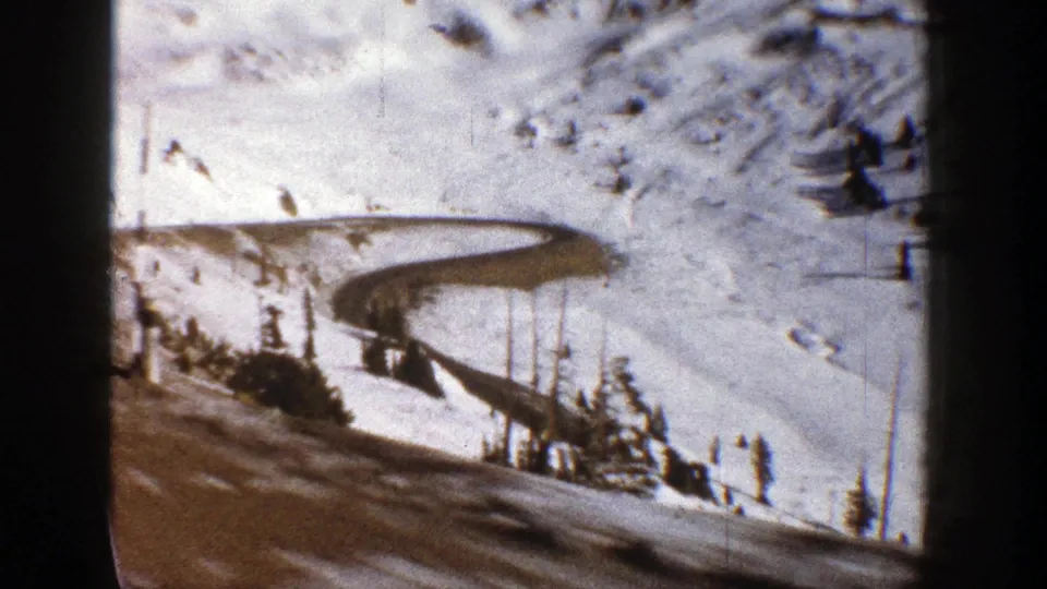 1961 Aspen winter road scene showing a curved mountain track, snow-covered hillside, evergreens, and interior vehicle framing.