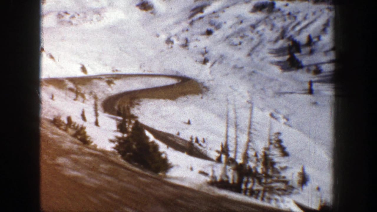 1961 Aspen 8mm frame showing a snow-covered slope, curving road, scattered evergreens, and exposed ground.