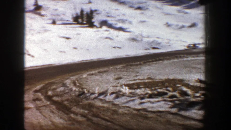 1961 Aspen 8mm frame looking across a snow-covered road curve with tracked snow and distant evergreens.