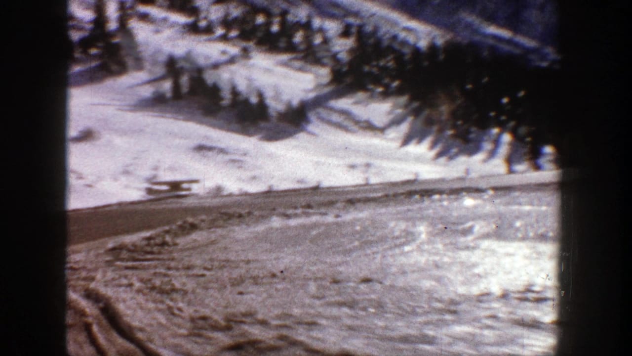 Snowy mountain road beside conifer-covered slopes, seen through dark frame edges.