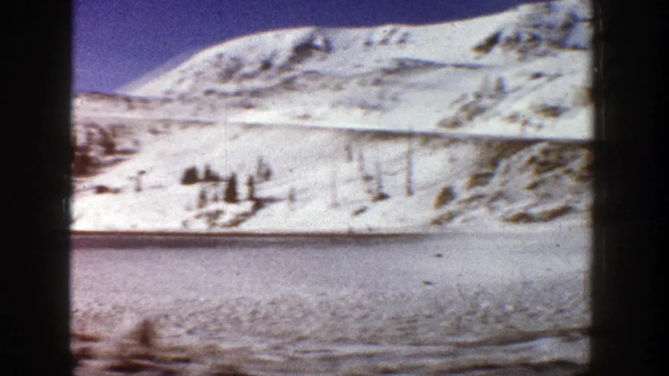 Aspen-area winter frame showing a snow-covered mountain slope, sparse conifers, and an icy roadside foreground on 8mm film.