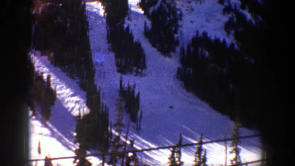 1961 home-movie frame of an Aspen ski slope descending between evergreen trees, with grain and dark gate edges.