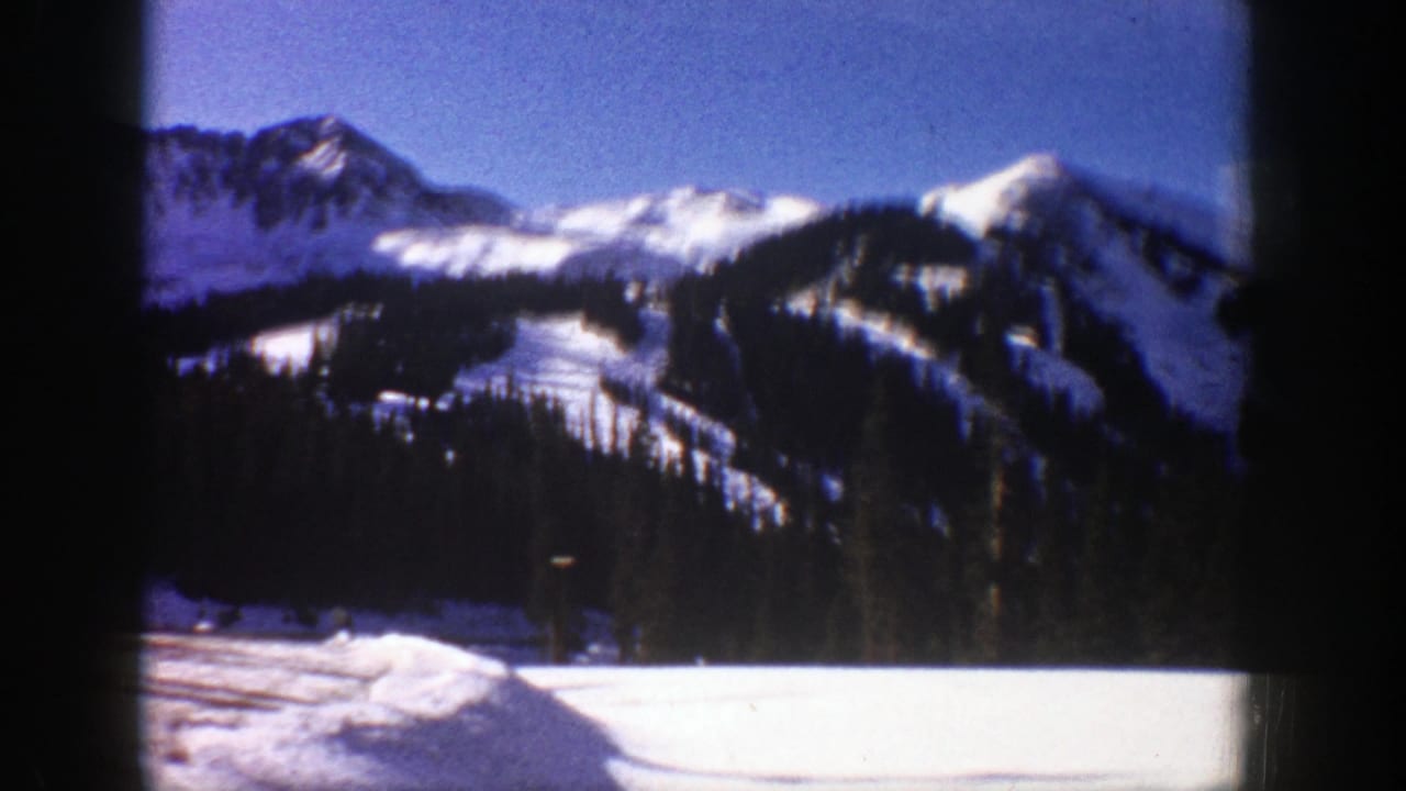 1961 Aspen-area winter mountain view with snow-covered slopes, dark pine forest, and blue sky on 8mm film.
