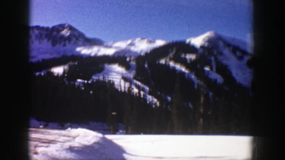 1961 Aspen-area winter mountain view with snow-covered slopes, dark pine forest, and blue sky on 8mm film.