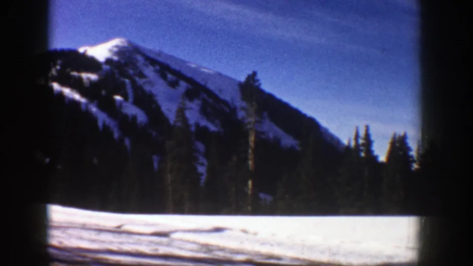 8mm home-movie frame of Aspen-area winter scenery, with snow, pines, and a mountain peak beneath a blue sky.